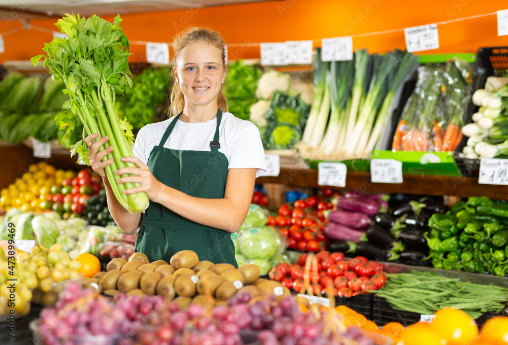 Portrait of positive teenage girl in uniform working in grocery shop as job experience, selling celery