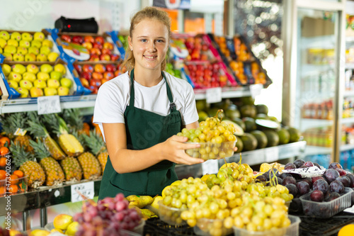 Obraz na plátně Hardworking fifteen-year-old girl who works part-time in a store as a trainee saleswoman puts grapes on the counter, laid
