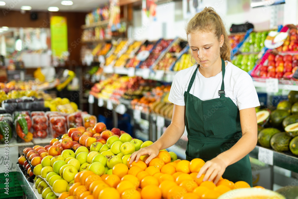Obraz premium Hardworking fifteen-year-old girl who works part-time in a store as a trainee saleswoman puts oranges on the counter
