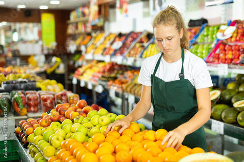 Hardworking fifteen-year-old girl who works part-time in a store as a trainee saleswoman puts oranges on the counter