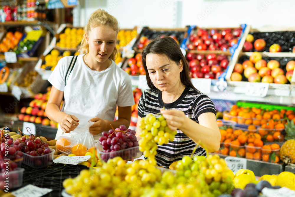 Portrait of teenage girl and her mother who buying fresh red grape at grocery shop