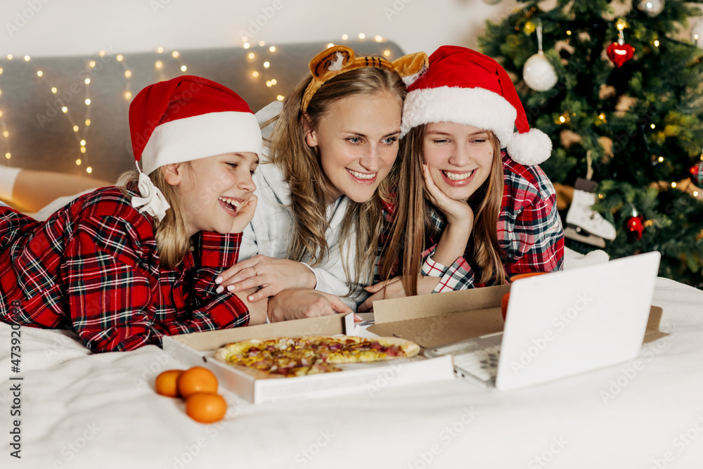 A young mother and her two wonderful daughters enjoy a pizza delivered and a movie in the evening, sitting on the bed on Christmas or New Year's Eve A young mother and her two wonderful daughters enjoy a pizza delivered and a movie in the evening, sitting on the bed on Christmas or New Year's Eve