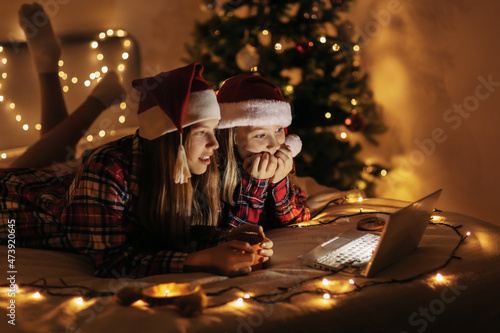Two teenage girls in plaid shirts lying on the bed and using a laptop, a lighted Christmas tree in the background. Christmas atmosphere, Christmas time, time of magic