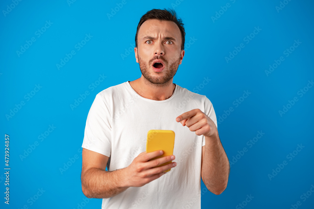 Astonished guy looking at smartphone screen in shock over blue background Astonished guy looking at smartphone screen in shock over blue background
