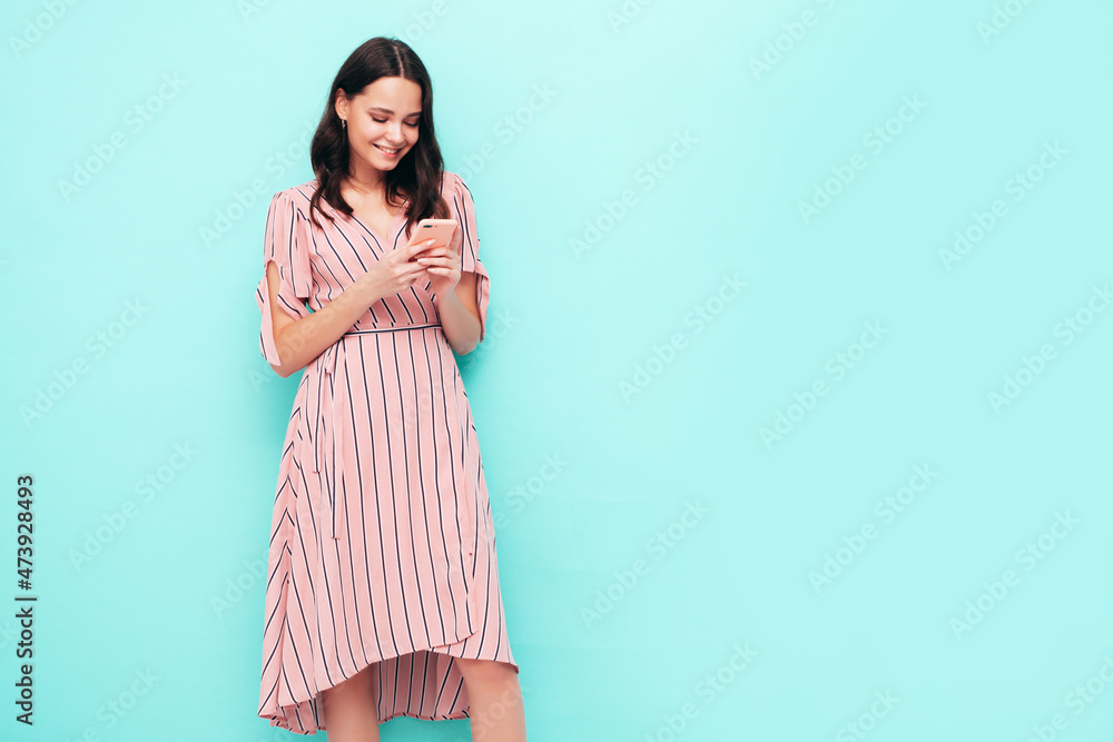 Young beautiful smiling female in trendy summer dress. Sexy carefree woman posing near blue wall in studio. Positive brunette model looking at cellphone screen. Holding smartphone and using apps Young beautiful smiling female in trendy summer dress. Sexy carefree woman posing near blue wall in studio. Positive brunette model looking at cellphone screen. Holding smartphone and using apps