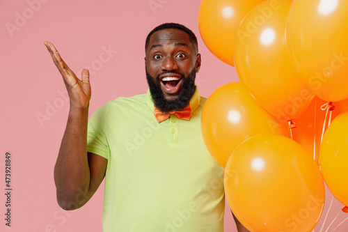 Young surprised black gay man 20s in green t-shirt hat cone hold bunch of air inflated helium balloons celebrating birthday party spread hands isolated on plain pastel pink background studio portrait © ViDi Studio