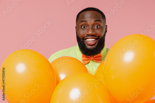 Young smiling happy fun cool black gay man 20s in green t-shirt bow tie hold bunch of air inflated helium balloons celebrating birthday party isolated on plain pastel pink background studio portrait © ViDi Studio