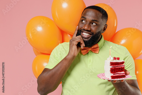 Young minded black gay man 20s in green t-shirt bow tie hold bunch of air inflated helium balloons celebrate birthday party sweet cake with candle look aside isolated on plain pastel pink background © ViDi Studio