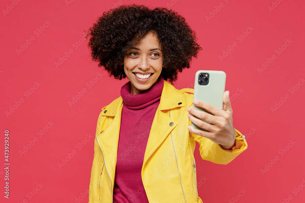 Charming fun happy young curly black latin woman 20s years old wears yellow jacket doing selfie shot on mobile cell phone post photo on social network isolated on plain red background studio portrait. Charming fun happy young curly black latin woman 20s years old wears yellow jacket doing selfie shot on mobile cell phone post photo on social network isolated on plain red background studio portrait.