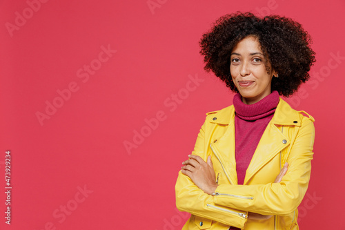 Charming bright young curly black latin woman 20s years old wears yellow jacket looking camera hold hands crossed isolated on plain red background studio portrait. People emotions lifestyle concept.
