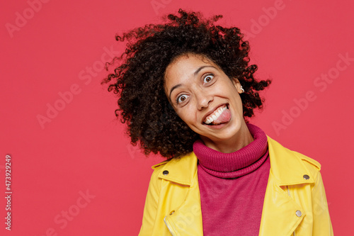 Young curly black latin woman 20s wears yellow jacket dance waving fooling around have fun enjoy play fluttering hair show make stick tongue out sign isolated on plain red background studio portrait.