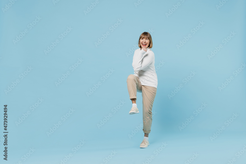 Full body elderly happy smiling woman 50s wearing white knitted sweater do winner gesture clench fist raise up leg isolated on plain blue color background studio portrait. People lifestyle concept Full body elderly happy smiling woman 50s wearing white knitted sweater do winner gesture clench fist raise up leg isolated on plain blue color background studio portrait. People lifestyle concept