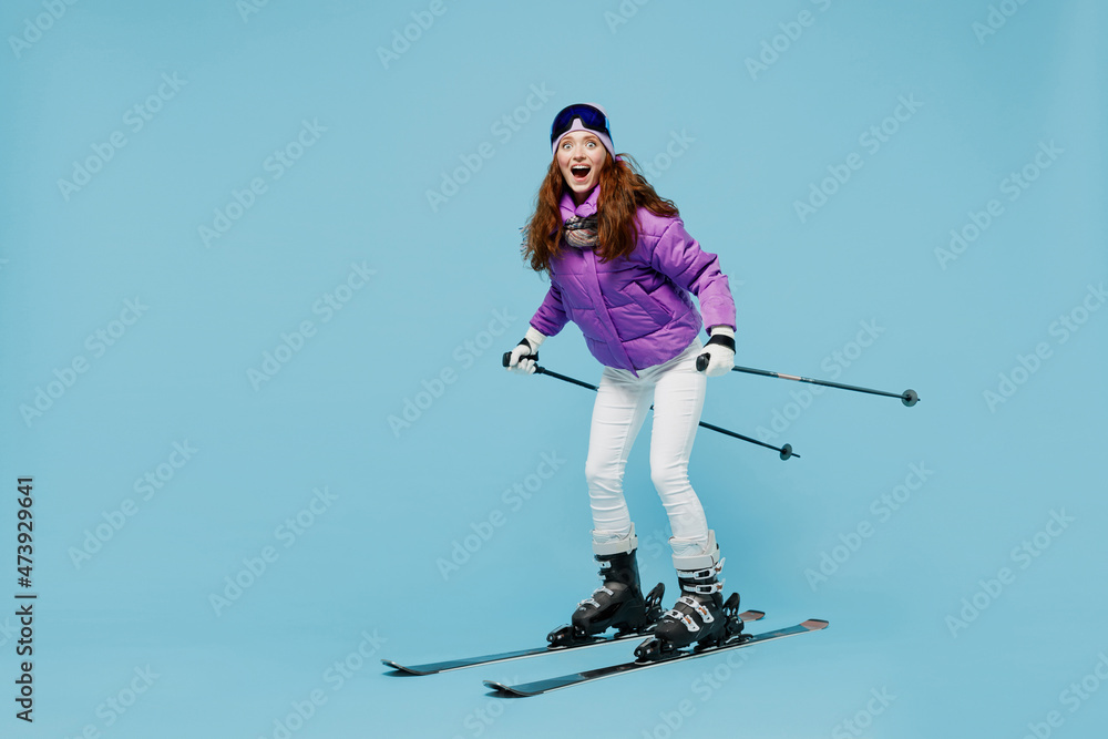 Full body skier amazed happy fun cool woman 20s wearing warm purple padded windbreaker jacket ski goggles mask spend extreme weekend in mountains look camera isolated on plain blue background studio