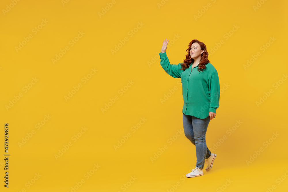 Full size body length side view young ginger chubby overweight woman 20s wears green shirt go move step meet greet waving hand as notices someone isolated on plain yellow background studio portrait.