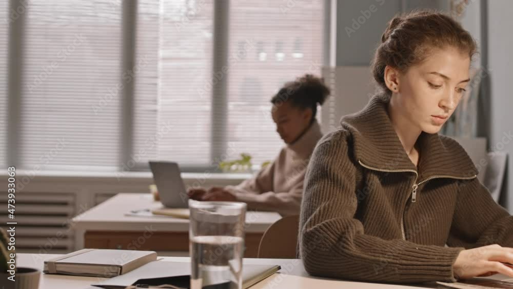 Tracking waist up slowmo of two young multiethnic women typing on laptops sitting at their workplaces in small cozy office Tracking waist up slowmo of two young multiethnic women typing on laptops sitting at their workplaces in small cozy office