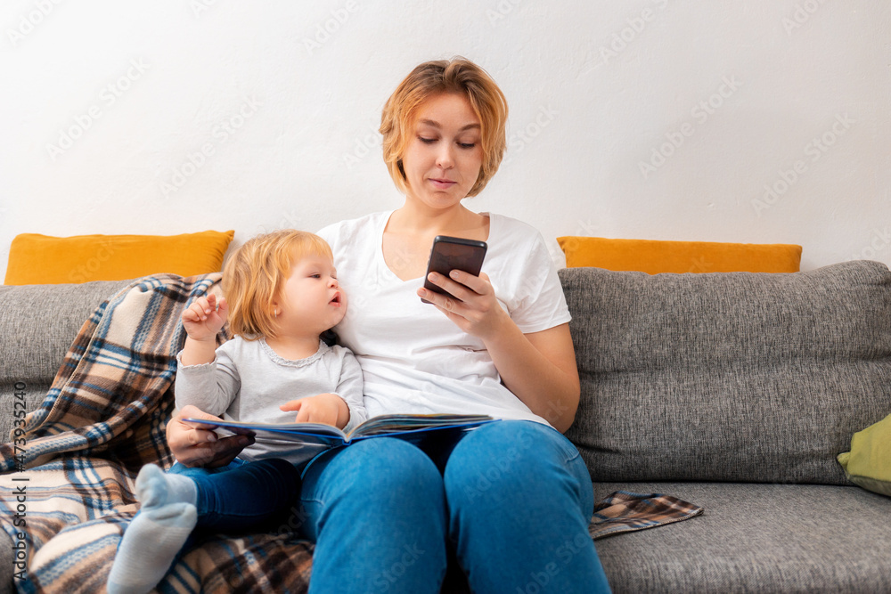 A mother sits with a little child on the couch and uses a smartphone. The kid looks into the smartphone. The concept of family relaxation