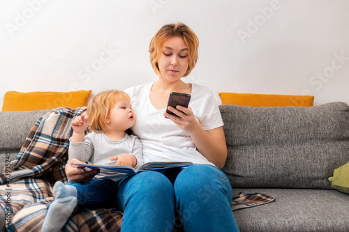 A mother sits with a little child on the couch and uses a smartphone. The kid looks into the smartphone. The concept of family relaxation