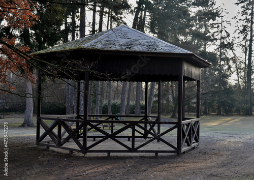 polygonal pergola with benches and a fence around the perimeter. there is a lot of space inside. the roof is pyramidal covered with asphalt shingles, in natural park, footpath, gravel © Michal