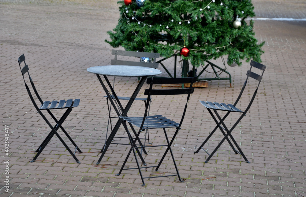metal black chairs and tables in a cafe on a cobbled square. everything is made of light material that can be folded and cleaned every night. Christmas tree with glass ornaments