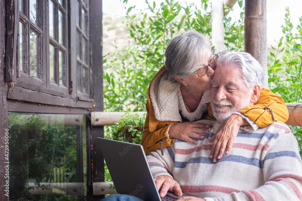 Carefree senior couple hugging and kissing in wooden balcony of mountain chalet while man using laptop computer. Carefree senior couple hugging and kissing in wooden balcony of mountain chalet while man using laptop computer.