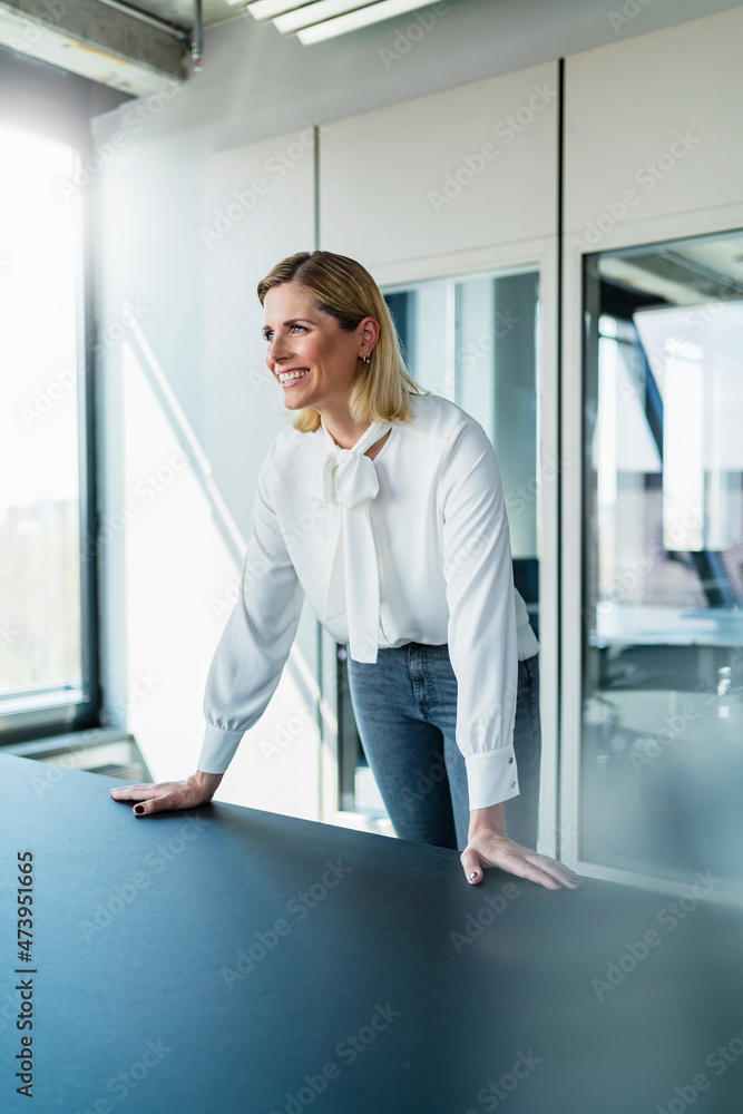Smiling blond businesswoman leaning on desk while standing in office looking away Smiling blond businesswoman leaning on desk while standing in office looking away