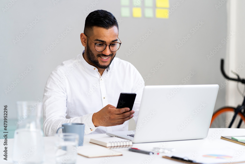 Young businessman using smart phone by laptop on desk at office Young businessman using smart phone by laptop on desk at office
