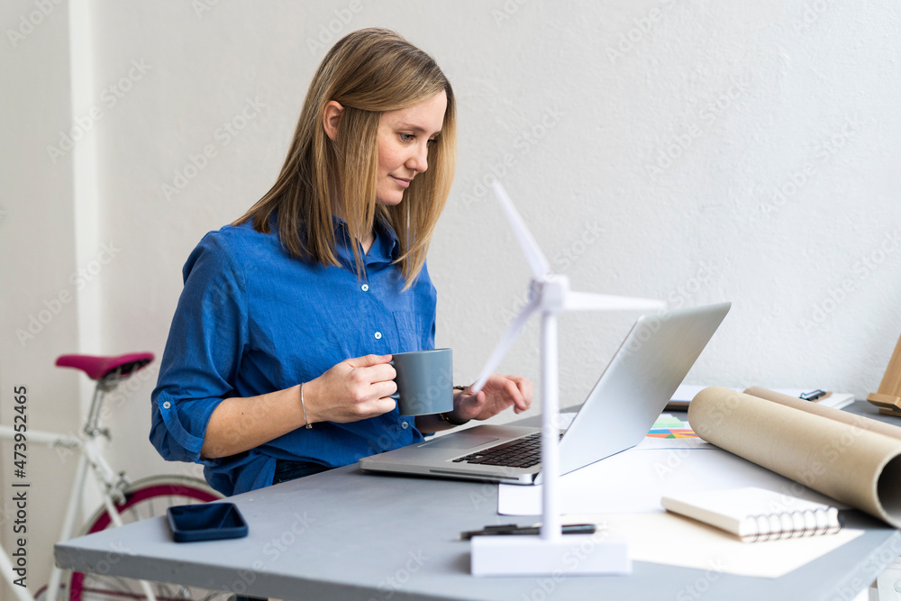 Businesswoman holding coffee cup while using laptop in office Businesswoman holding coffee cup while using laptop in office