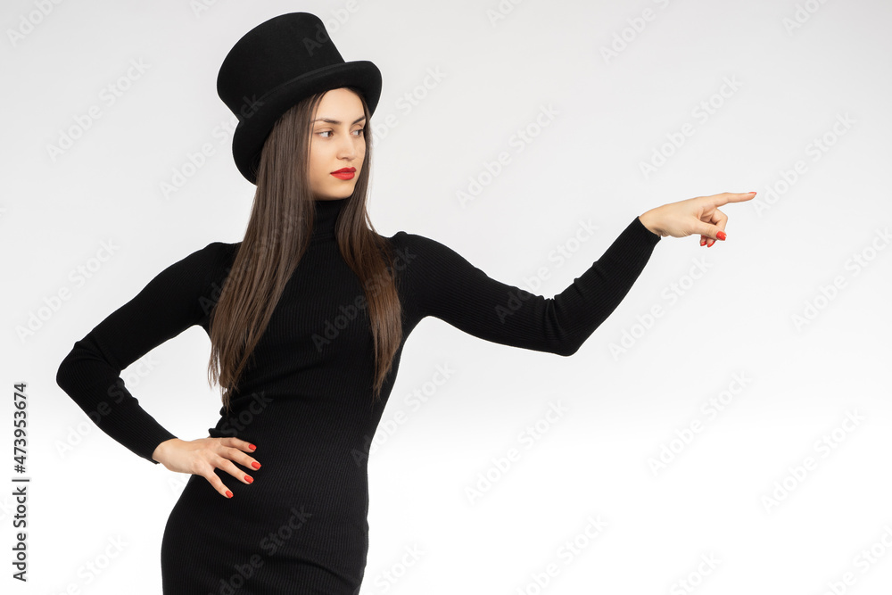 Girl in black clothes points her finger at something. Place for an advertising message. Woman in a black hat and dress. A long-haired girl draws attention to some information. Female in black dress. Girl in black clothes points her finger at something. Place for an advertising message. Woman in a black hat and dress. A long-haired girl draws attention to some information. Female in black dress.