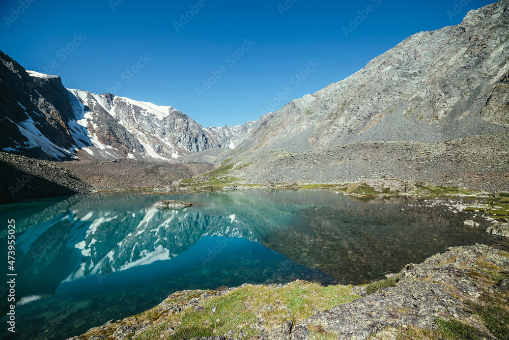 Fototapeta premium Snowy mountain reflected in clear water of glacial lake. Beautiful sunny landscape with glacier reflection in water surface of mountain lake under clear sky. Snow on rock reflected in mountain lake.