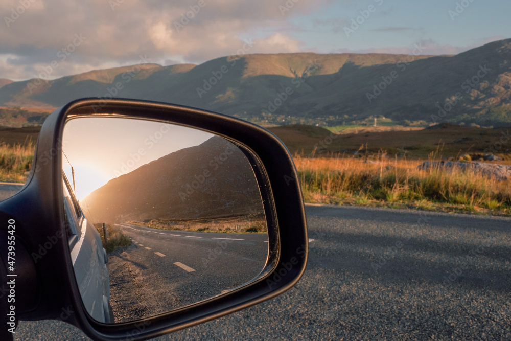 Road and sun rise view in a car mirror, stunning mountains out of focus in the background. Connemara, county Galway, Ireland. Car travel and tourism concept. Road and sun rise view in a car mirror, stunning mountains out of focus in the background. Connemara, county Galway, Ireland. Car travel and tourism concept.