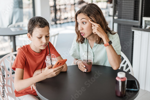 Emotional conflict between mother and son about smartphone addiction in cafe. An angry mom and a teenager ignoring her.