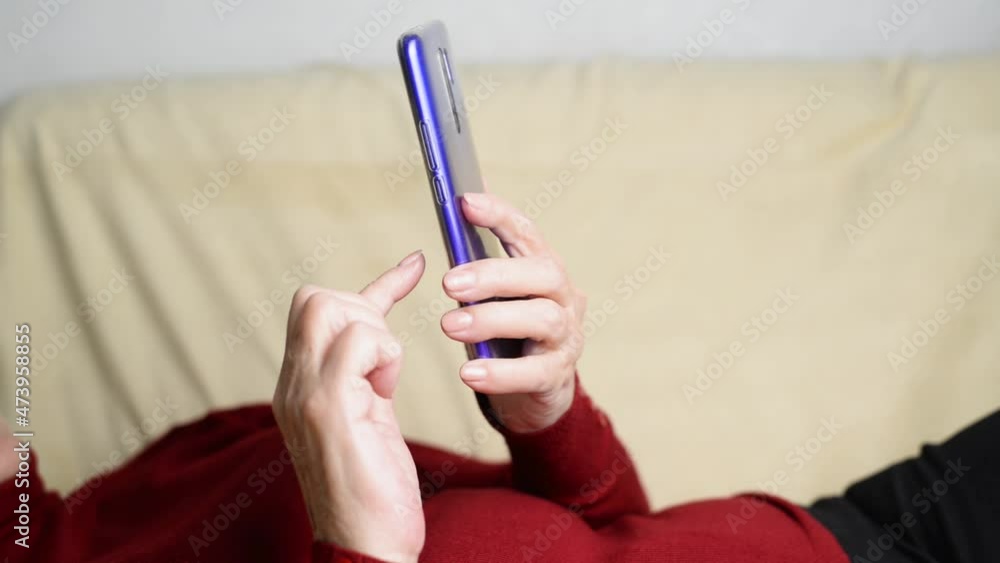 Side view of relaxing senior woman using mobile phone, scrolling, surfing internet touching smartphone screen while lying on couch at home. Elderly and technology. Selective focus on phone Side view of relaxing senior woman using mobile phone, scrolling, surfing internet touching smartphone screen while lying on couch at home. Elderly and technology. Selective focus on phone