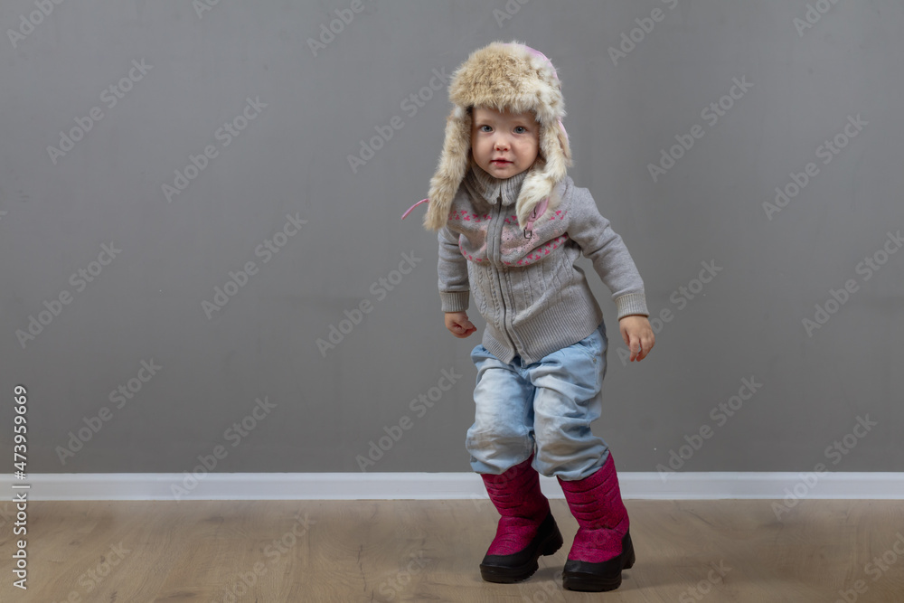 Cute baby girl 2 years old caucasian in warm winter clothes and a hat with earflaps with fur on a gray background Cute baby girl 2 years old caucasian in warm winter clothes and a hat with earflaps with fur on a gray background
