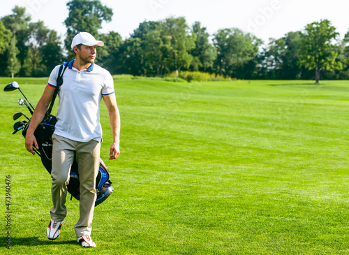 Young Male Golfer carries a Bag of Golf Clubs. Slim Man in White T-shirt , Golf Shoes, White Cap and Beige Pants is Going in the Green Turf Grass. Copy Space. Full-length