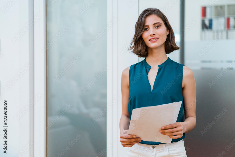 Beautiful young female entrepreneur with documents standing at doorway in office Beautiful young female entrepreneur with documents standing at doorway in office