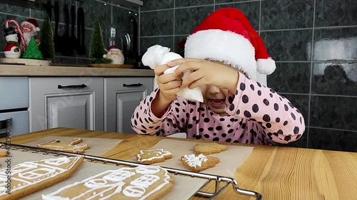 Merry Christmas and Happy Holidays. Little girl in Santa hat thinking how to decorate a cookie decorates gingerbread cookies using glaze.