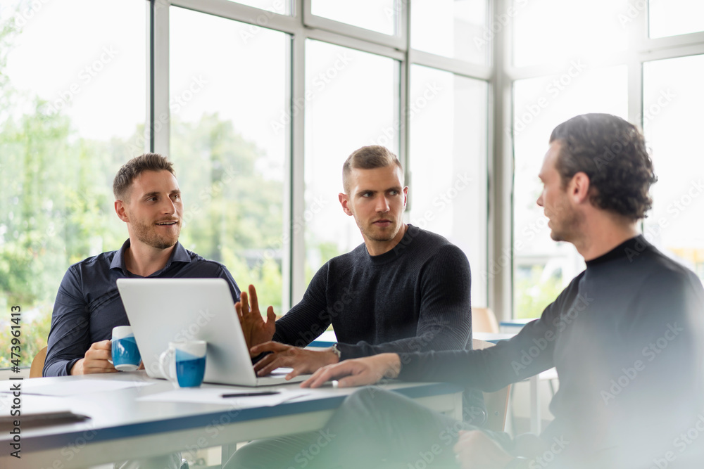 Male professionals sitting with laptop while discussing at desk in office Male professionals sitting with laptop while discussing at desk in office