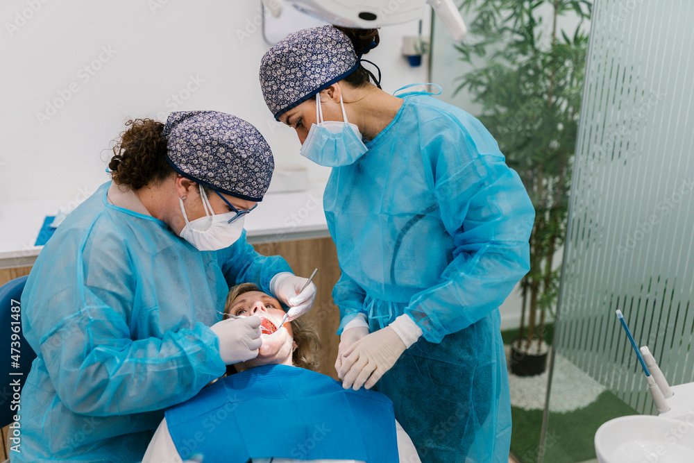 Orthodontists in protective workwear and face mask checking patient while standing by assistant in clinic Orthodontists in protective workwear and face mask checking patient while standing by assistant in clinic