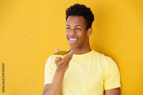Smiling man making voice note through smart phone in front of yellow wall