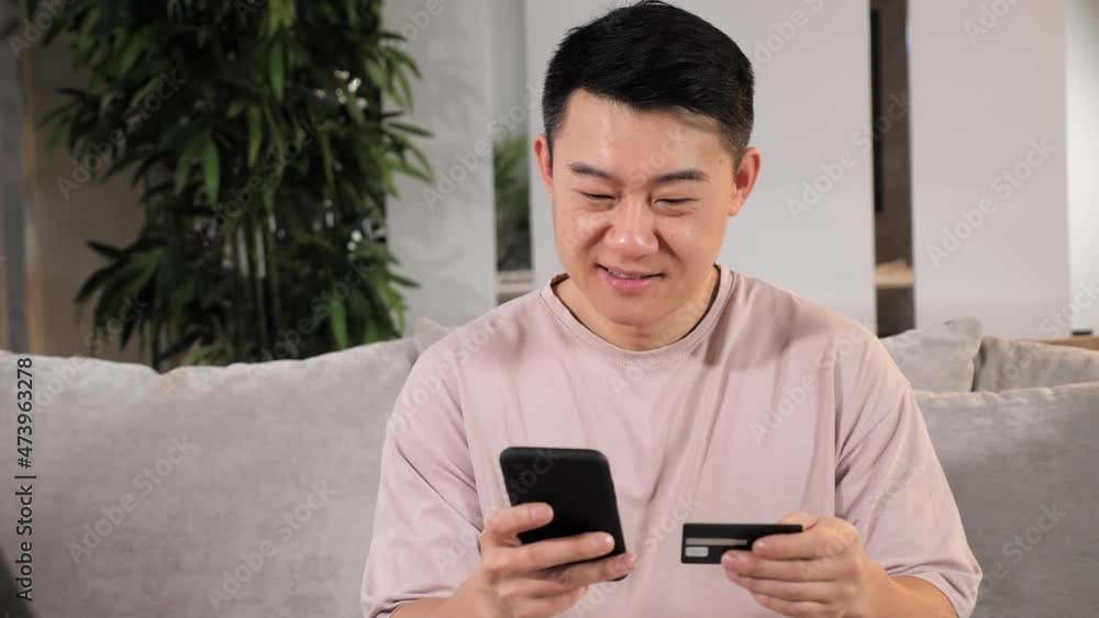 Finance shopping in internet. Smiling asian man customer holding credit card smartphone sitting on couch at home. Happy male shopper using instant easy mobile payments making purchase in online store. Finance shopping in internet. Smiling asian man customer holding credit card smartphone sitting on couch at home. Happy male shopper using instant easy mobile payments making purchase in online store.