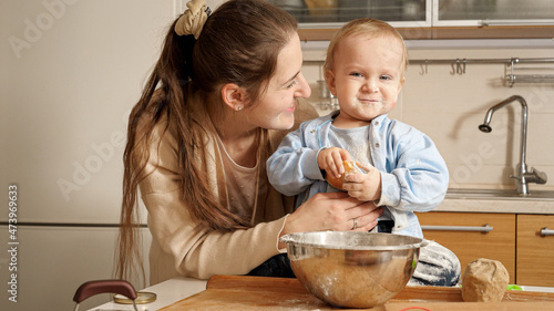 Smiling young mother with little baby son making bread dough in bowl on kitchen. Concept of little chef, children cooking food, healthy nutrition.
