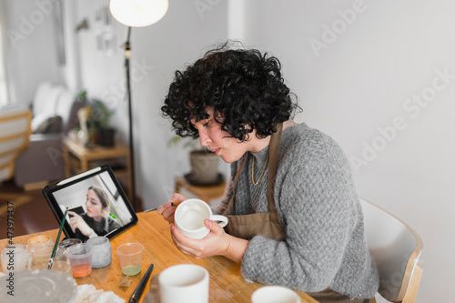 Artist painting cup while learning through online tutorial on digital tablet at workplace
