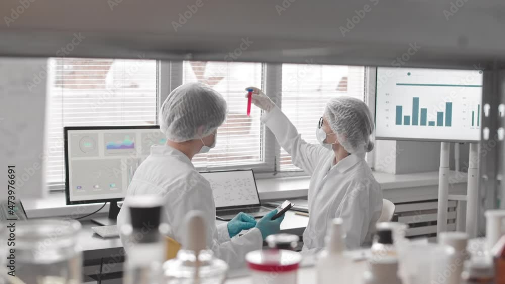 Medium of two Caucasian female scientists wearing face masks, gloves, medical gowns and hats sitting at desk in laboratory at daytime, examining test tube, talking and using tablet computer Medium of two Caucasian female scientists wearing face masks, gloves, medical gowns and hats sitting at desk in laboratory at daytime, examining test tube, talking and using tablet computer