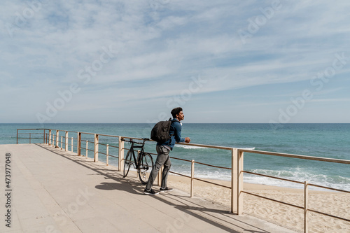 Mid adult man carrying backpack while leaning on railing by bicycle at beach during sunny day photo
