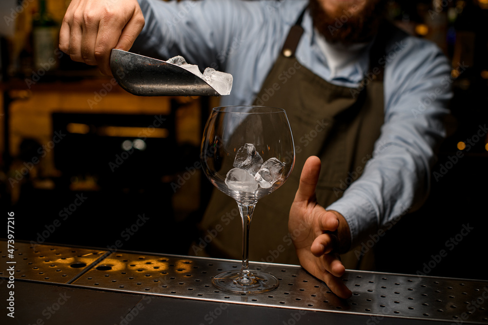 transparent wine goblet glass in which bartender pours ice cubes from a scoop