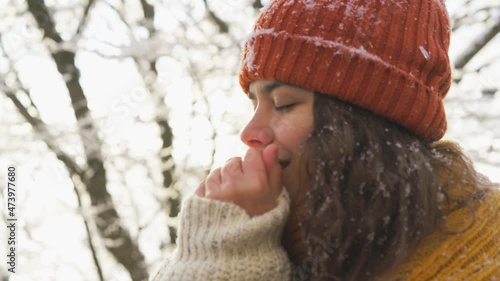 Close-up portrait woman in hat and scarf over sweater enjoys winter forest. Warms hands by rubbing palms and smiles looking at the camera standing against trees with frozen branches 