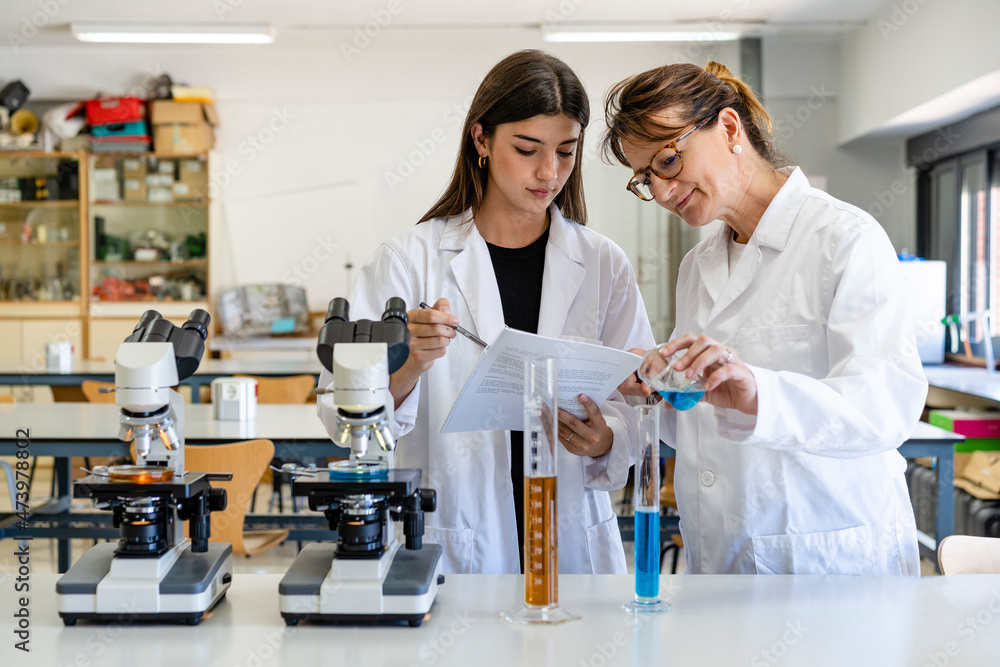 Female scientist reading document while colleague pouring liquid in graduated cylinder Female scientist reading document while colleague pouring liquid in graduated cylinder