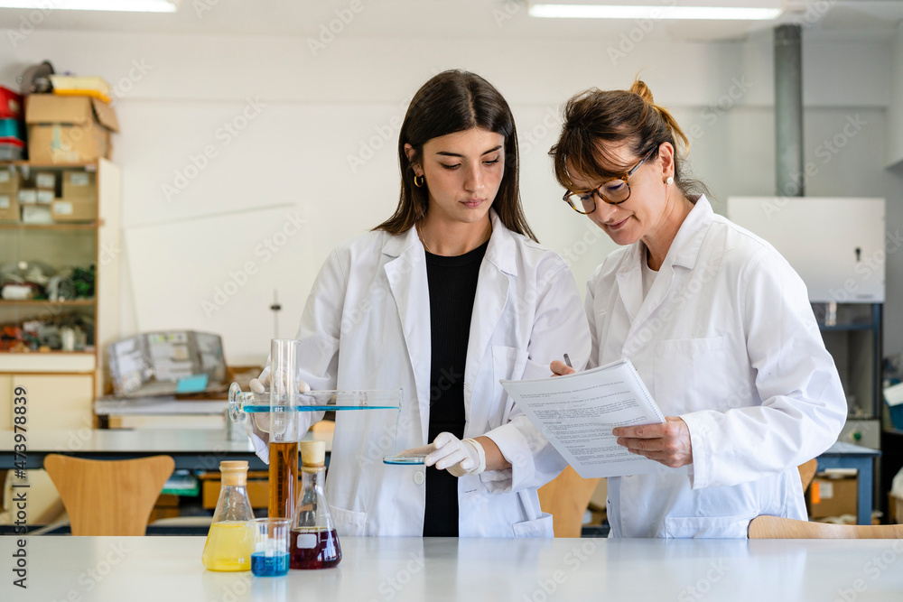 Female scientist reading document while assisting colleague experimenting in laboratory Female scientist reading document while assisting colleague experimenting in laboratory