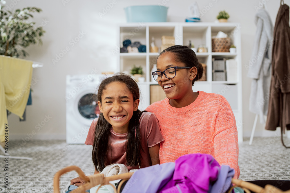 Obraz premium Smiling woman wearing glasses is sitting with pretty daughter on floor of bathroom laundry room in front of them wicker basket with colorful clothes in background dryer hangers washing machine