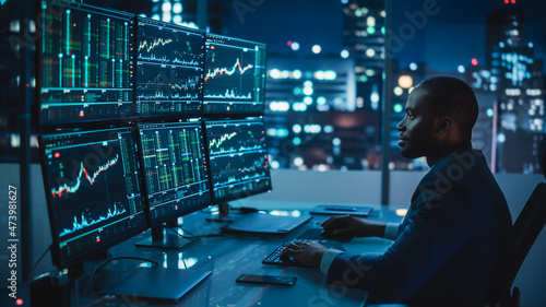 Financial Analyst Working on Computer with Multi-Monitor Workstation with Real-Time Stocks, Commodities and Exchange Market Charts. African American Trader Works in Investment Bank Late at Night.